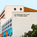 UT Health Austin sign on the south side of the Health Transformation Building as seen from the street with alternating flags of teal and burnt orange on the west side.