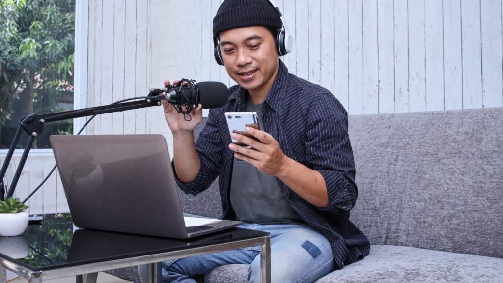A man is sitting on a couch, recording a voiceover while holding a cell phone in his hand.
