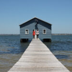 Fun Fact House on the water with a long dock and a couple holding hands in the distance.