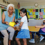 An older adult woman helps a young girl with schoolwork in a classroom, surrounded by other students.