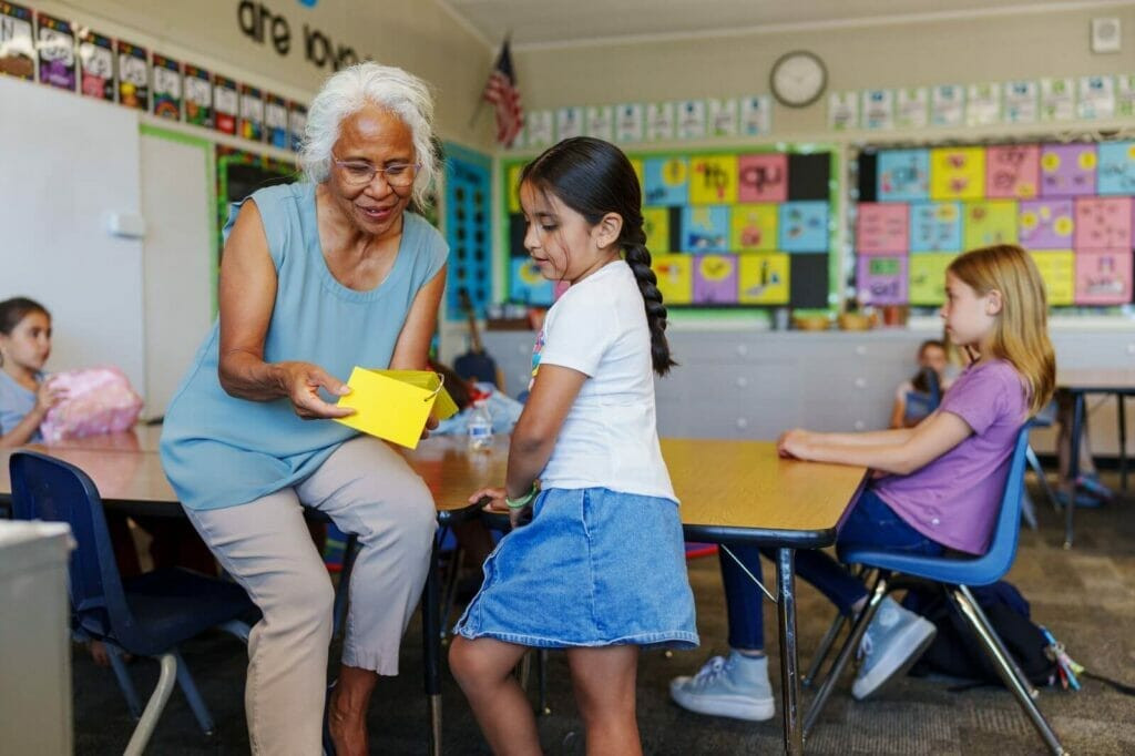 An older adult woman helps a young girl with schoolwork in a classroom, surrounded by other students.
