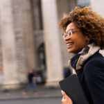 A Black woman smiles as she walks down the street holding a laptop case.